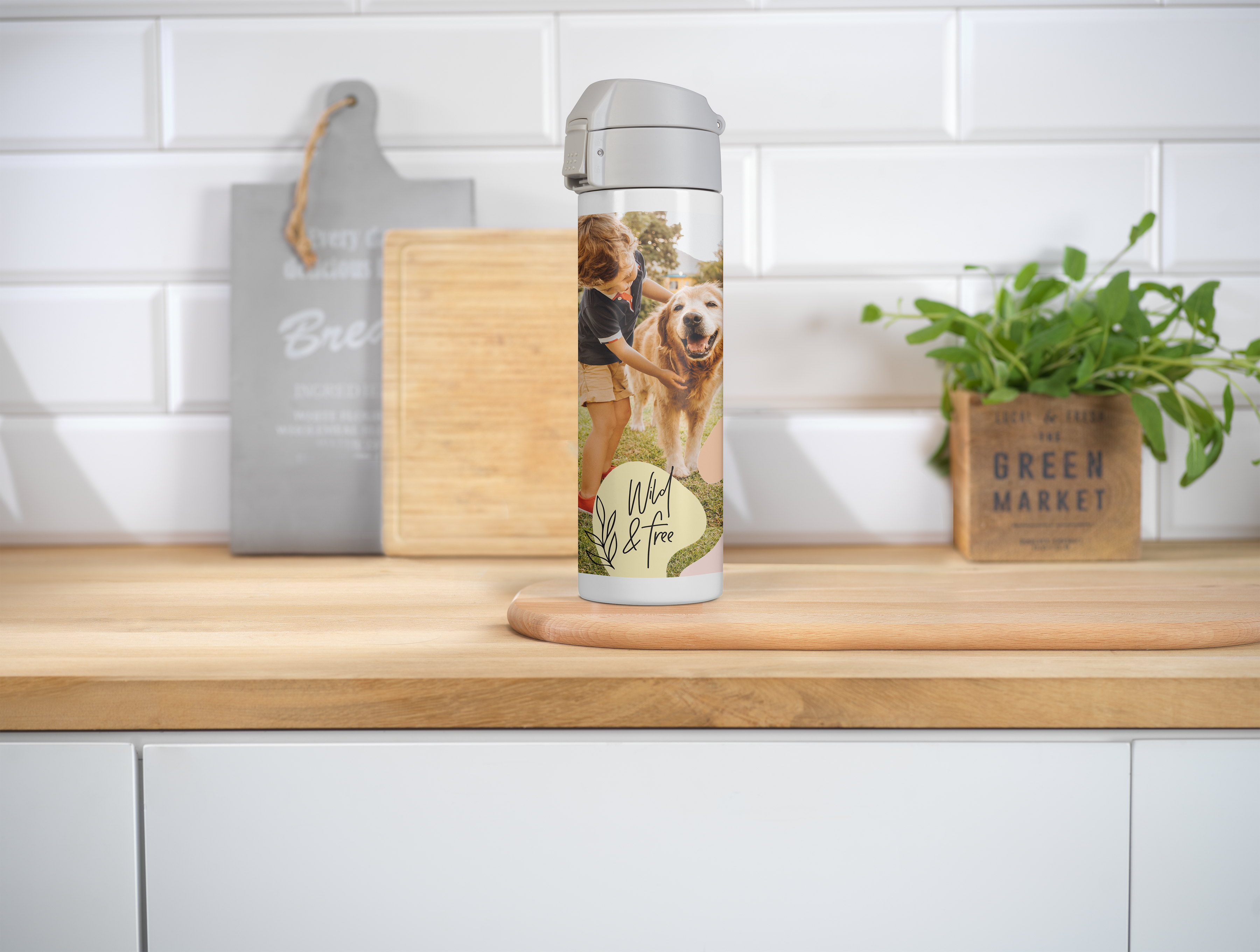 Een roestvrijstalen thermosfles met een foto van een kind en een hond, staat op een houten aanrecht in een moderne keuken met witte tegels en groene planten.