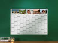 Pixum Jahresplaner Ein Jahresplaner für das Jahr 2024 hängt an einer grünen Wand, geschmückt mit verschiedenen saisonalen Fotos über den Monatsnamen.