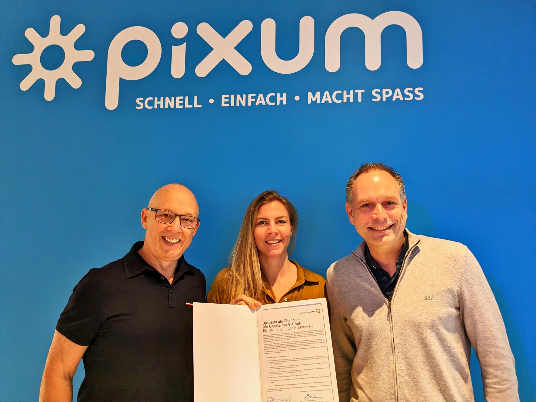 Daniel Attallah, Nora Engels und Marc Rendel mit der unterzeichneten Charta der Vielfalt vor dem Pixum Logo