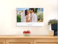 Fotoleinwand mit individuellem Text Eine Leinwand gestaltet mit Wunschtext und Foto hängt an einer . Auf der Leinwand ist ein Hochzeitsfoto eines Paares zu sehen und die Beschriftung "Emma & Leo - 15.07.2024".