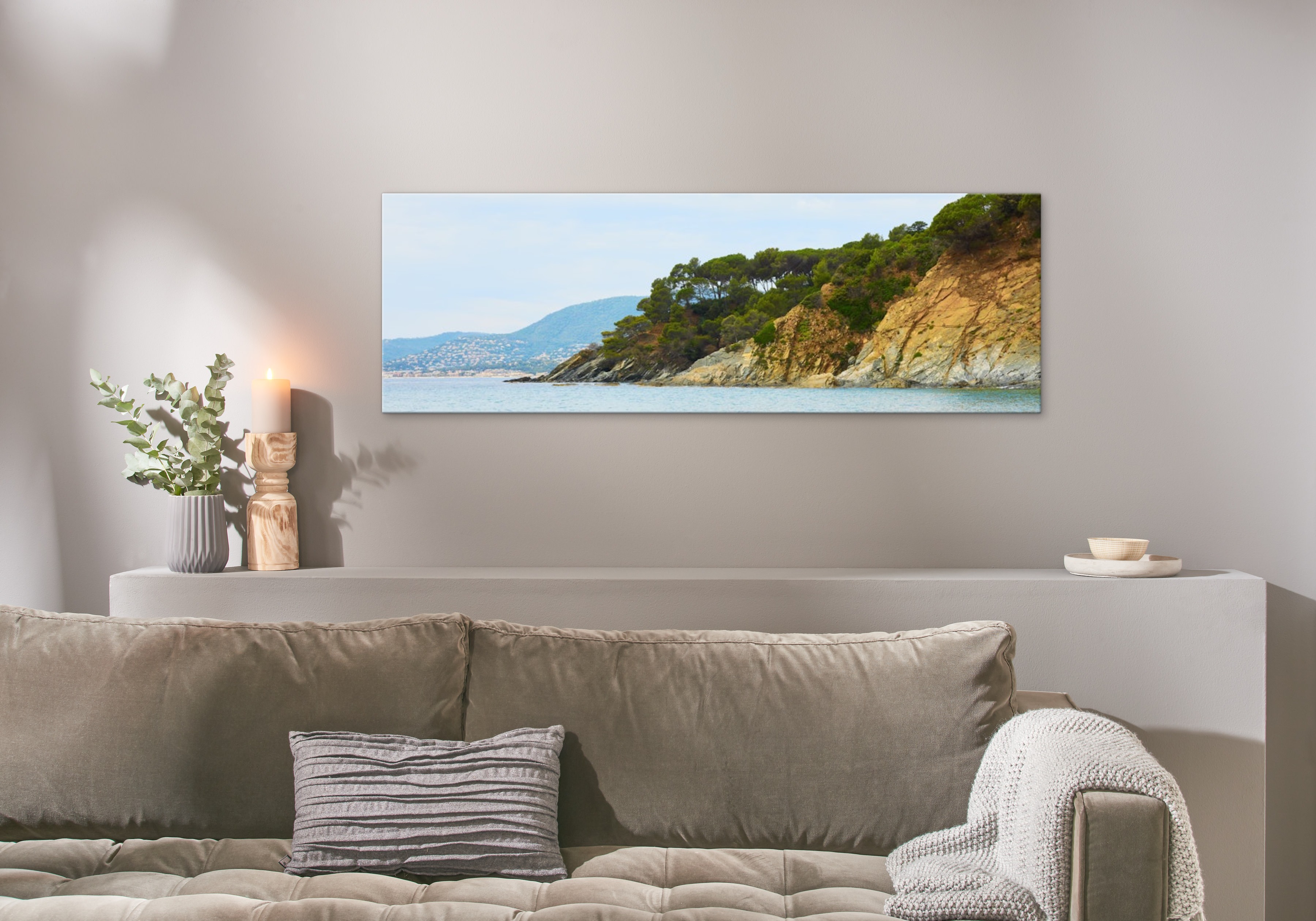 Eine Fotoleinwand im Format 150x50 cm hängt über einem Sofa und zeigt ein Bild von einer Küste.