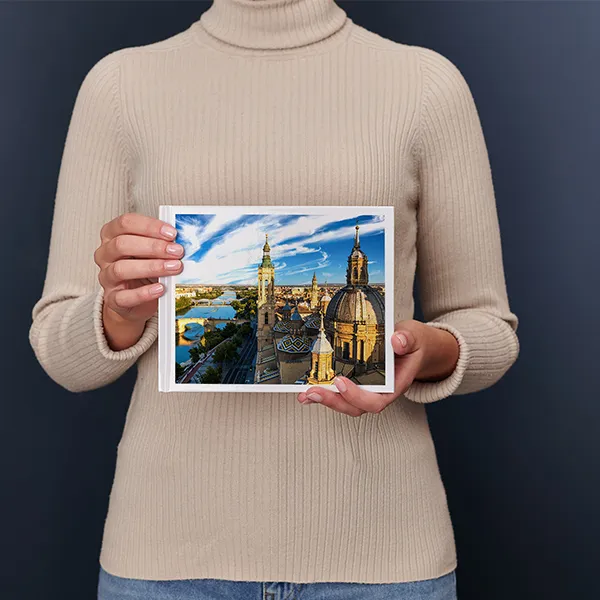 Een persoon in een beige trui houdt een fotoboek vast met daarop een lavendelveld met een boom en een huisje bij zonsondergang.