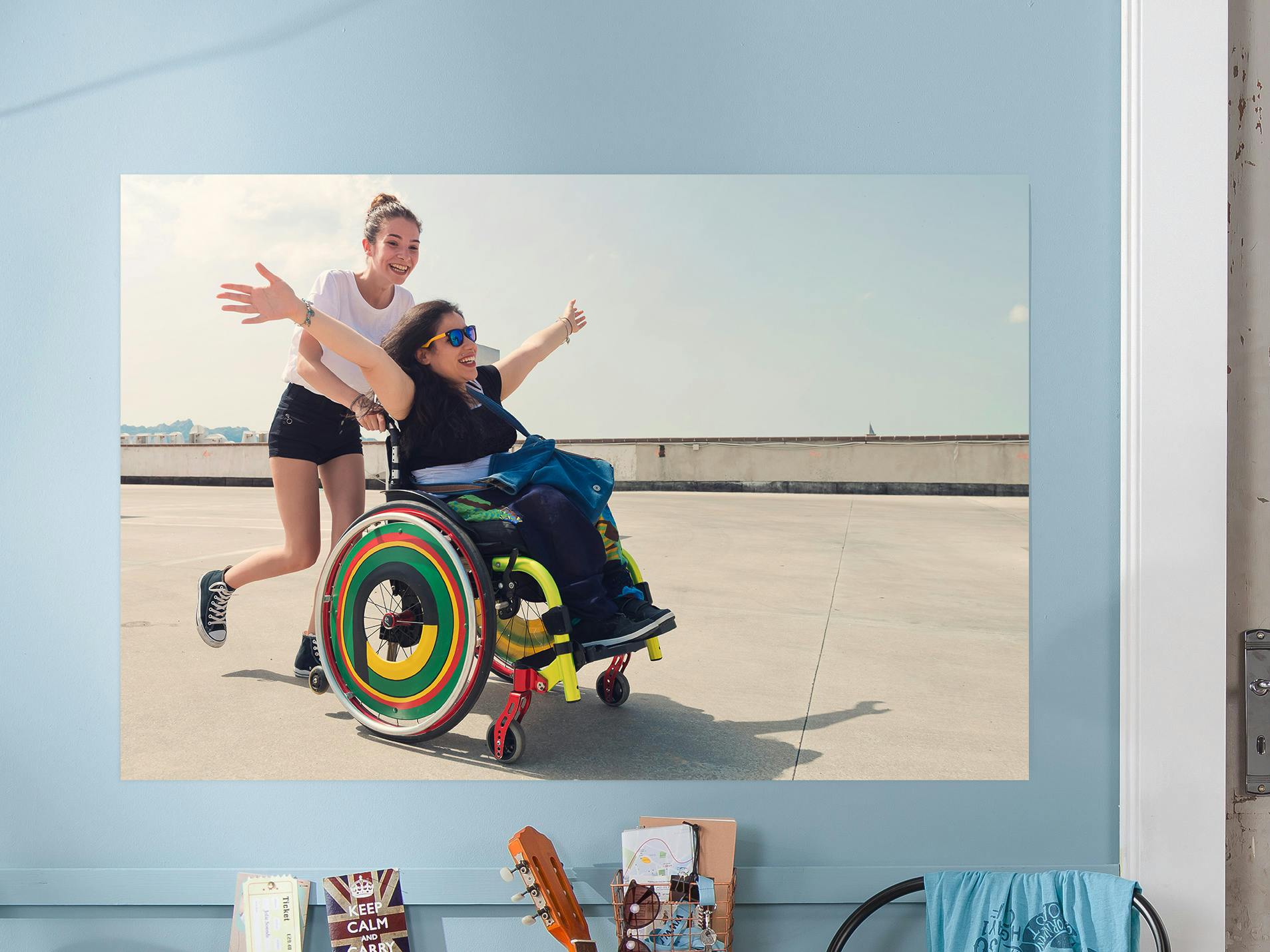 Fotoplakat med et motiv af to grinende venner, den ene sidder i en kørestol og den anden skubber til den