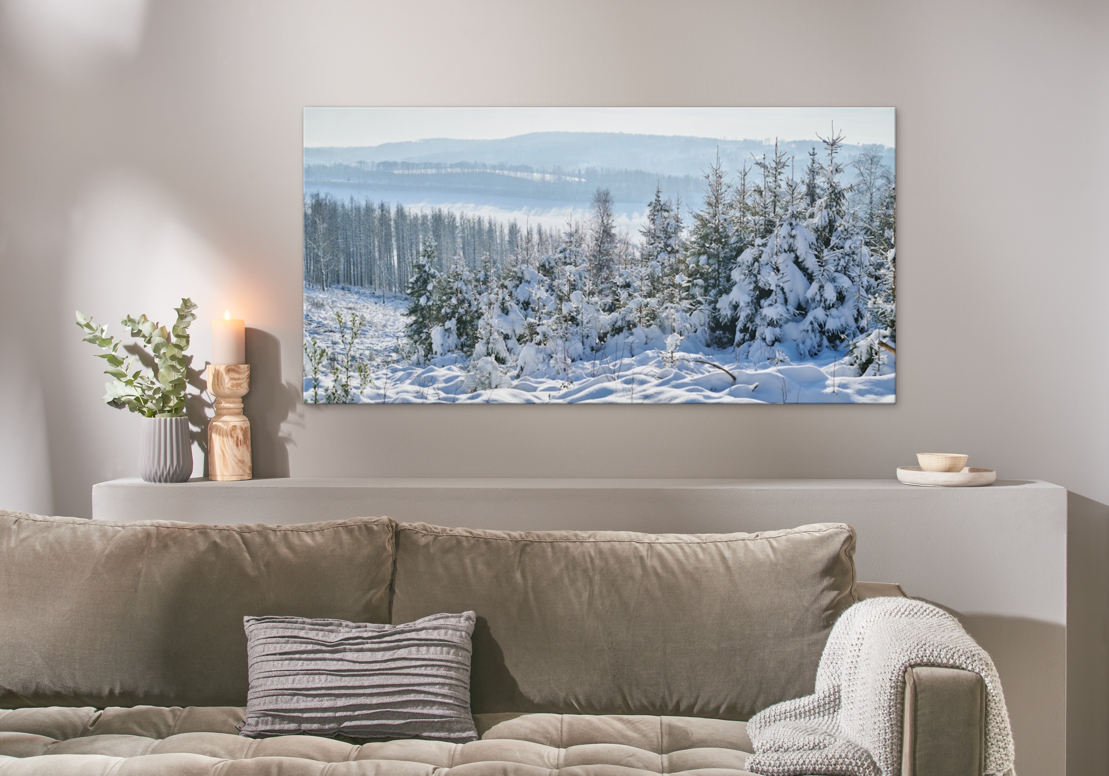 Eine Fotoleinwand im Format 160x80 cm hängt über einem Sofa und zeigt eine winterliche Wald-Landschaft.