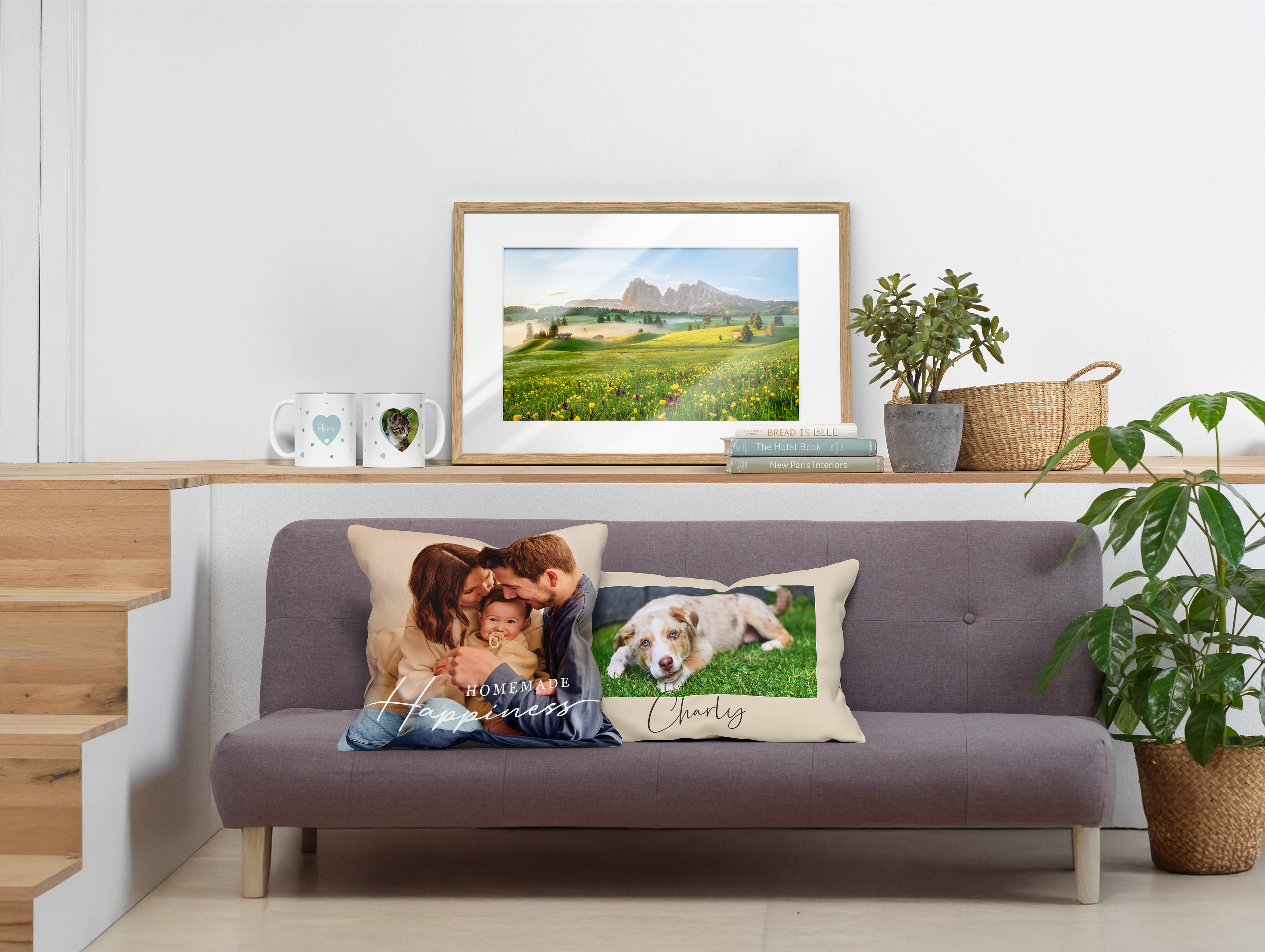 Modernes, helles Wohnzimmer mit grauem Sofa, dekoriert mit zwei personalisierten Fotokissen – eines zeigt ein Paar und das andere einen Hund auf einer Wiese mit dem Namen "Charly" darunter. Auf dem Regal über dem Sofa steht ein gerahmtes Landschaftsbild mit Wiese und Bergen, daneben Pflanzen, Bücher und zwei Tassen mit Herzmotiven. Auf der linken Seite führen Holzstufen nach oben, rechts steht eine große grüne Pflanze im Korb auf dem Boden.