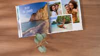 Pixum Fotobuch mit Urlaubsbildern Ein aufgeschlagenes Fotobuch zeigt links eine felsige Küste mit der Aufschrift 'Enjoy the Summer' und rechts eine Fotocollage mit einem Paar, einem tropischen Strand, einem Langboot und Sommermotiven.