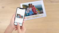 Álbum Digital en la app Una mano con un smartphone en el que se ha abierto la aplicación Pixum. Detrás hay un álbum de fotos Pixum con una foto de vacaciones en la portada.
