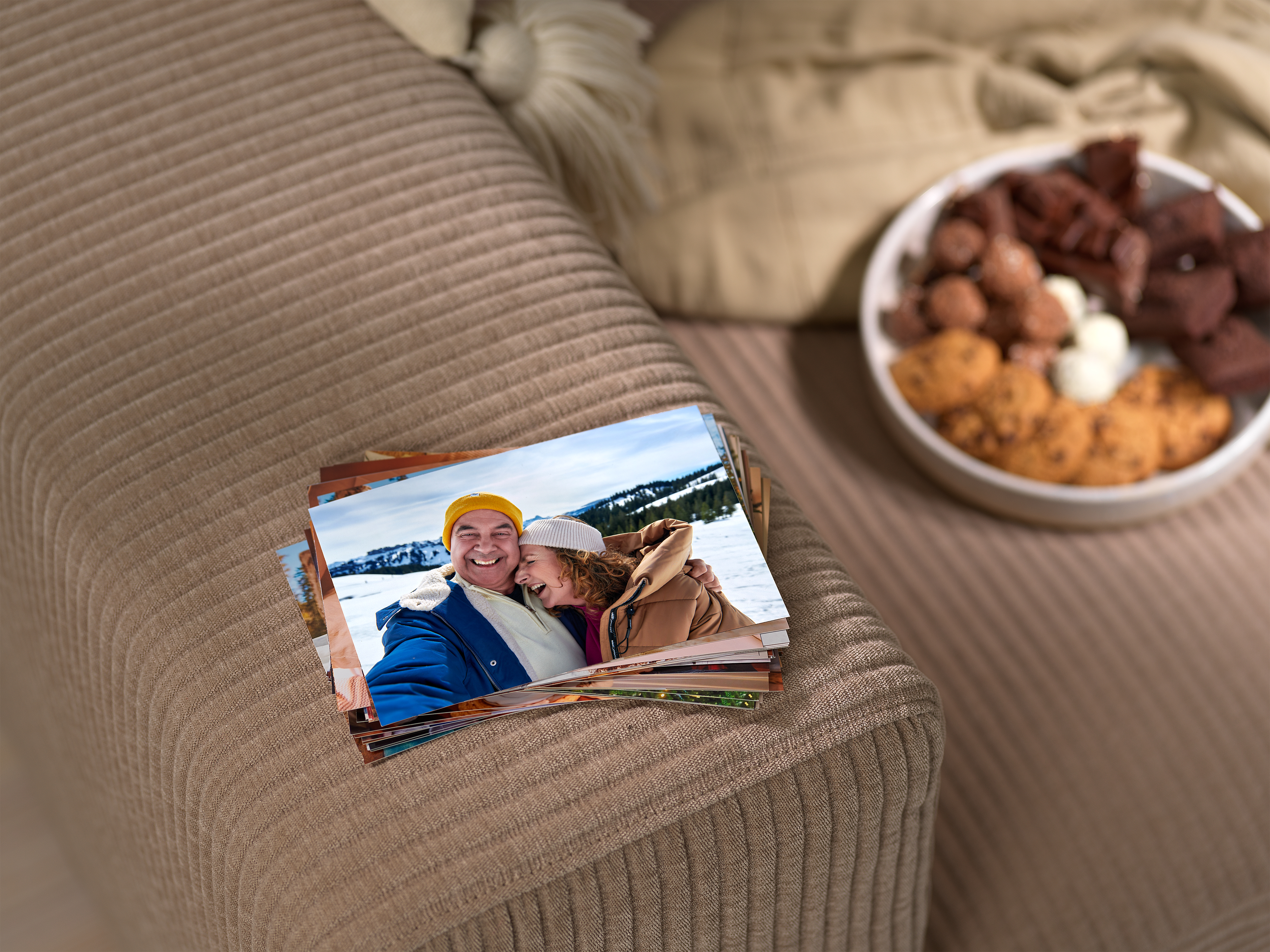 Ein Stapel von Fotoabzügen liegt auf einer beigen Couch. Auf den Abzügen sind Familienfotos abgebildet.