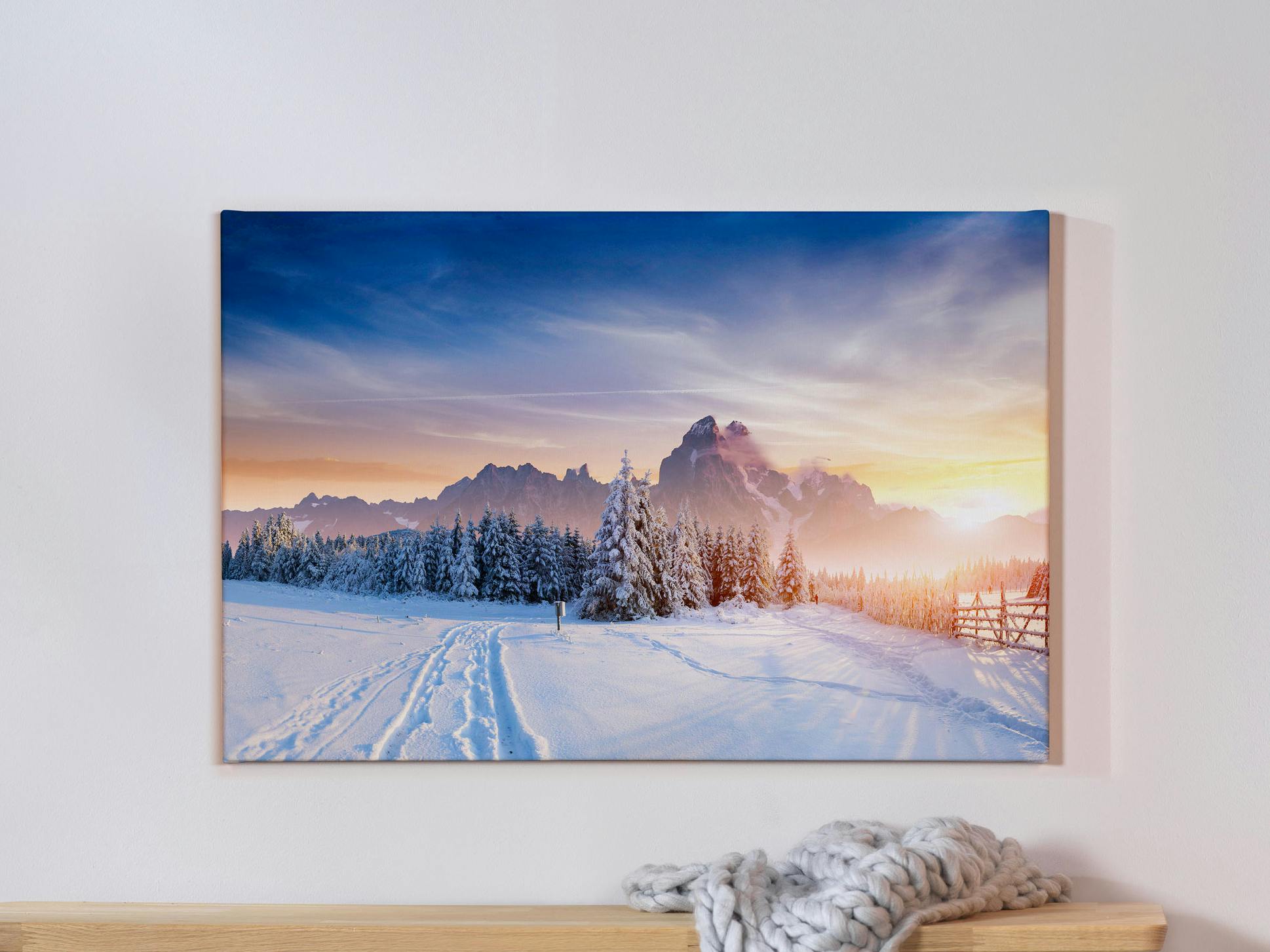 Fotoleinwand mit winterlichem Landschaftsmotiv auf einer grauen Wand und einer davorstehenden Bank mit einer Wolldecke