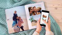 Fotobuch mit Cliparts per App gestalten Aufgeschlagenes Fotobuch im Hochformat mit sommerlichen Motiven und Cliparts und zwei Hände, die ein Smartphone halten