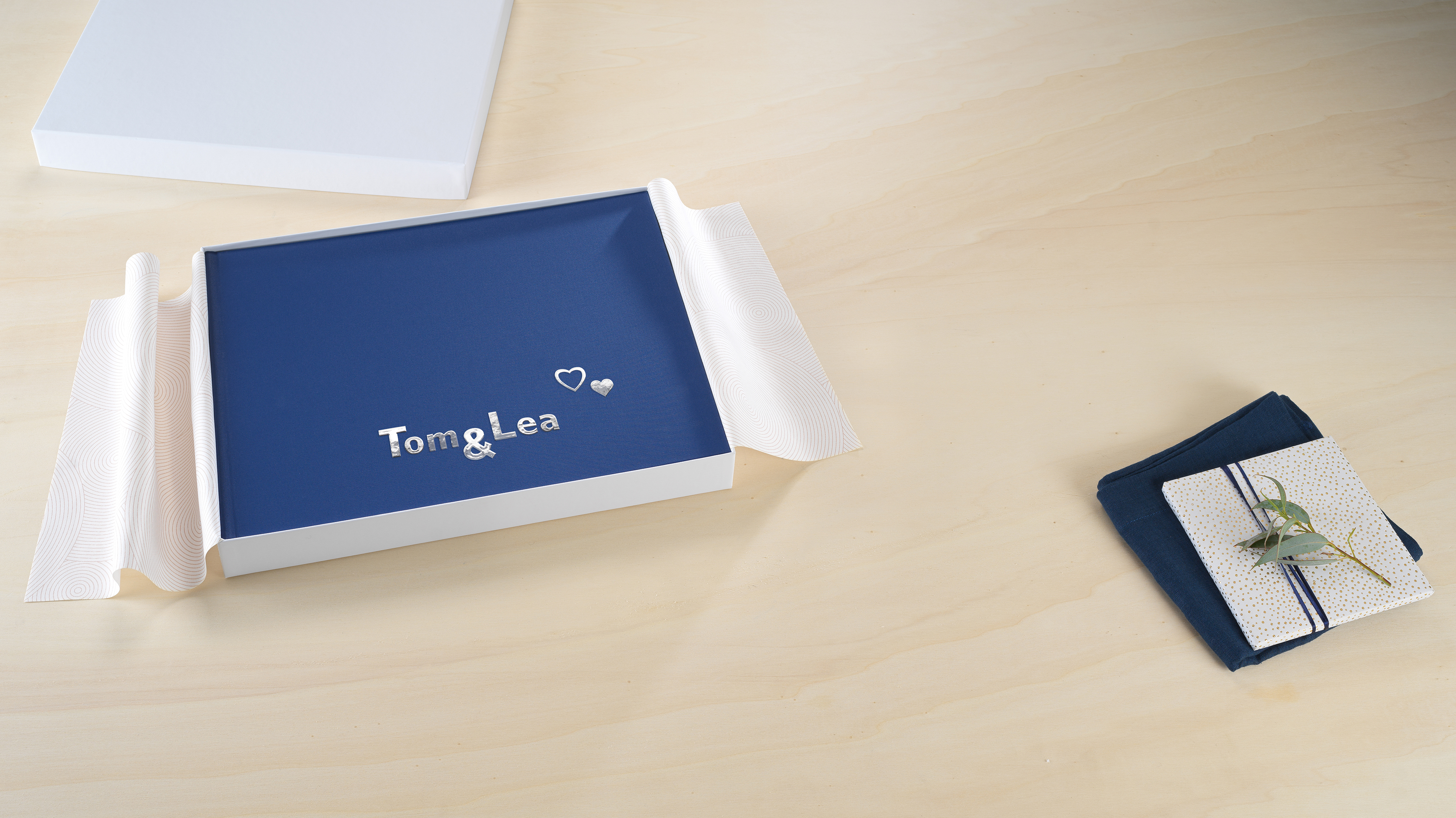 Een fotoboek met de tekst "Tom & Lea"  in een witte doos op een houten tafel, naast een gevouwen doek en een klein boekje met een takje erop.