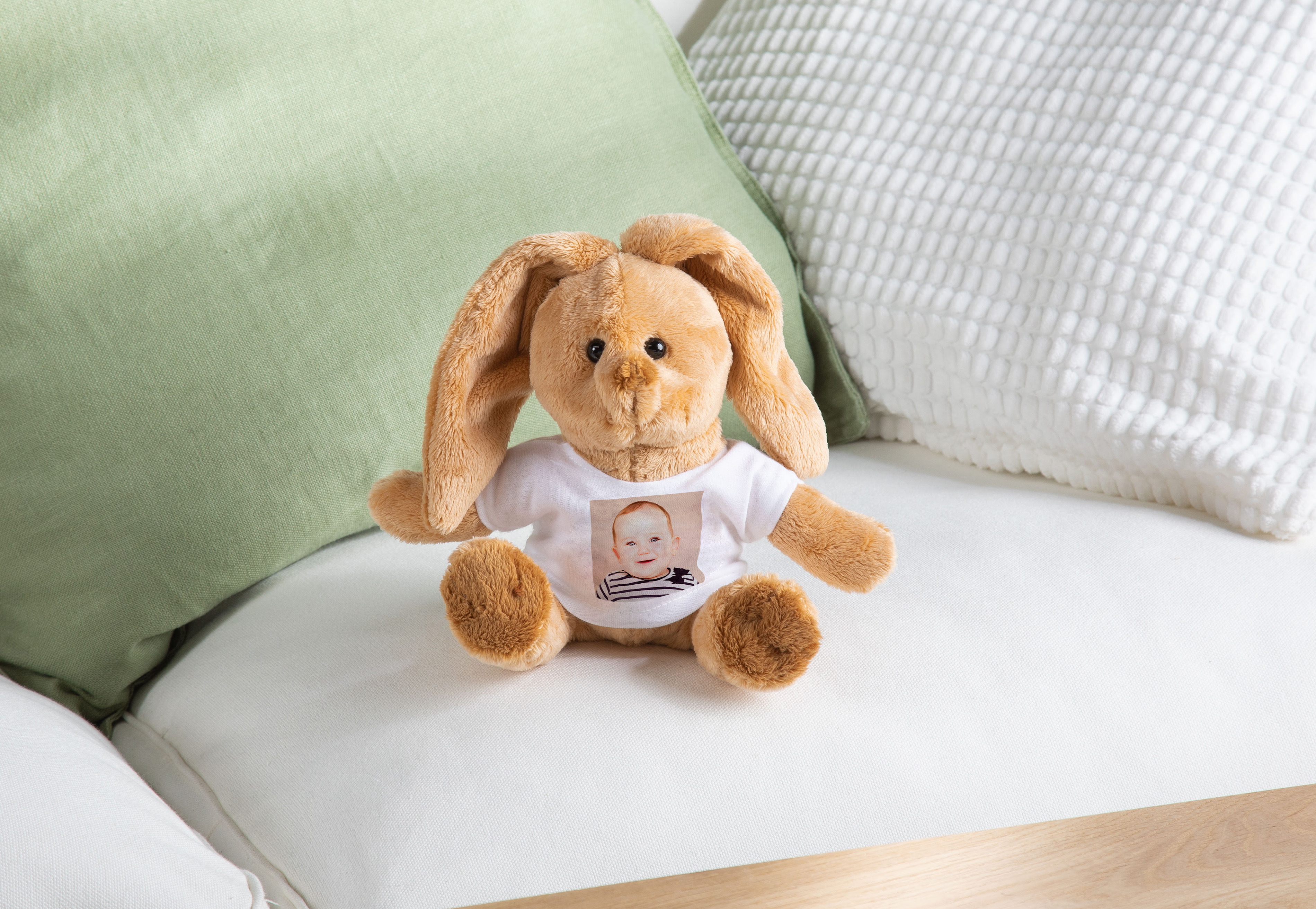 Ein brauner Plüschhase sitzt auf einem weißen Sofa. Der Hase trägt ein weißes T-Shirt, das mit einem Foto eines lächelnden Babys bedruckt ist.