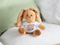 Ein brauner Plüschhase sitzt auf einem weißen Sofa. Der Hase trägt ein weißes T-Shirt, das mit einem Foto eines lächelnden Babys bedruckt ist.
