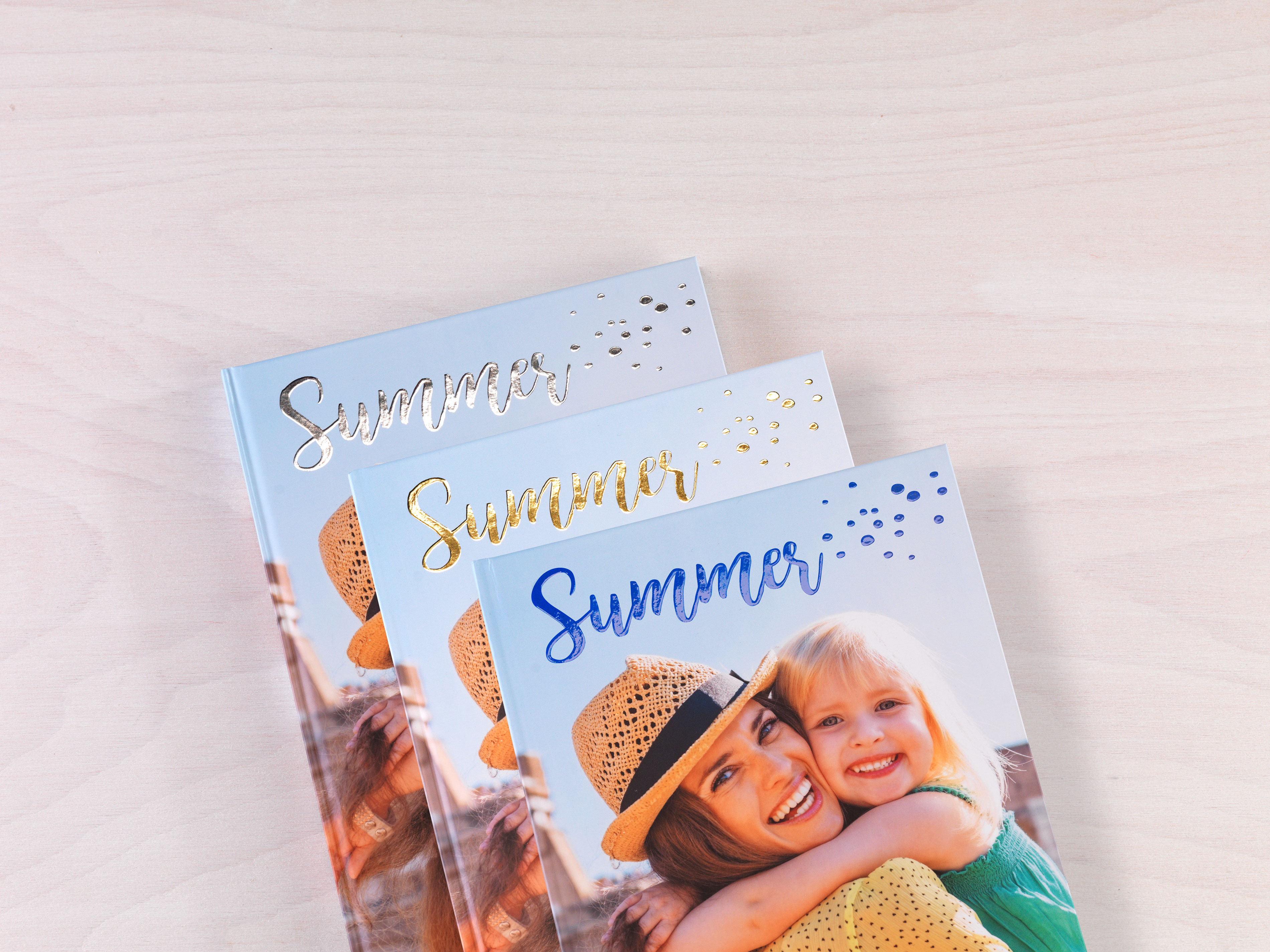 Drie fotoboeken met zomerthema liggen op elkaar; het bovenste toont een glimlachende vrouw en kind in zomerkleding.