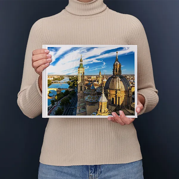 Een persoon houdt een fotoboek vast met daarop een lavendelveld met een huis en een boom, onder een kleurrijke avondlucht.