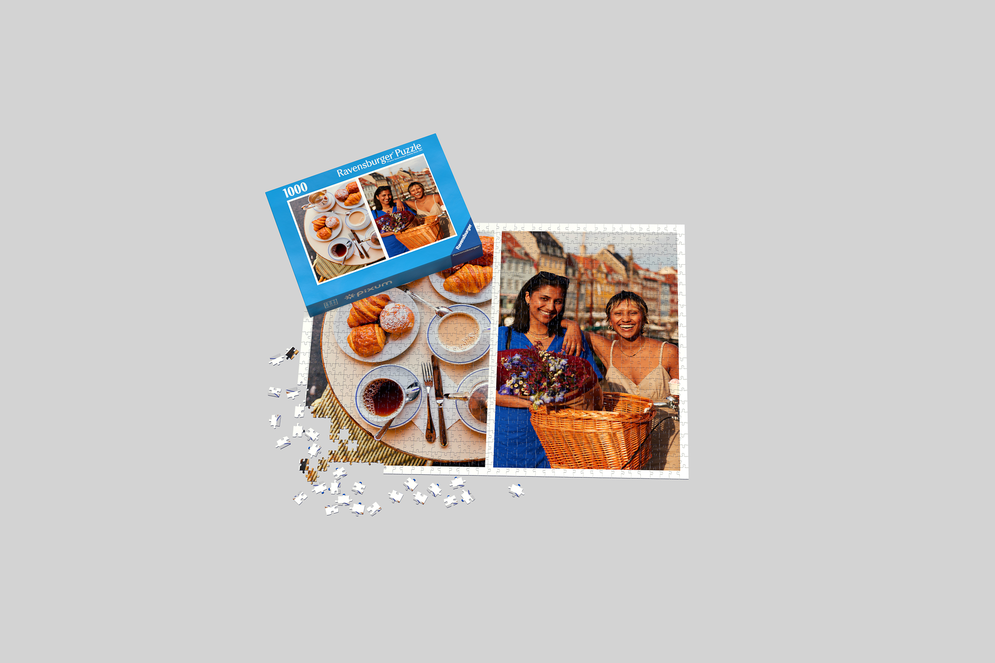 Auf einem Fotopuzzle mit 1000 Teilen sind zwei Fotos zu sehen. Auf dem einen stehen zwei Frauen mit einem Fahrrad vor einer Häuserreihe. Das andere Foto zeigt ein Frühstück mit Café und Croissants. Neben dem Fotopuzzle liegt die Box.