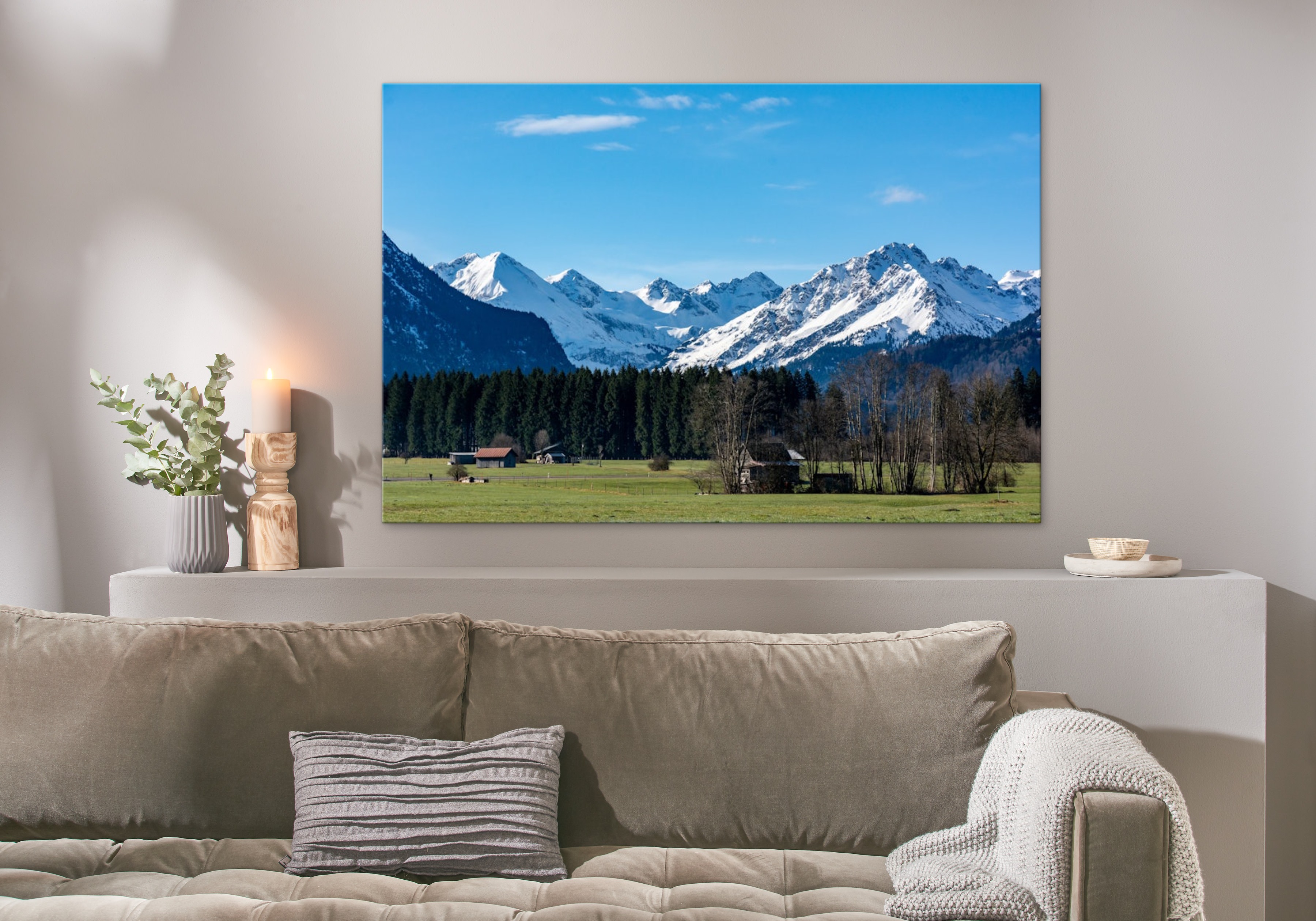 Eine Fotoleinwand im Format 150x100 cm hängt über einem Sofa und zeigt ein Bild mit Bergen und grüner Wiese.
