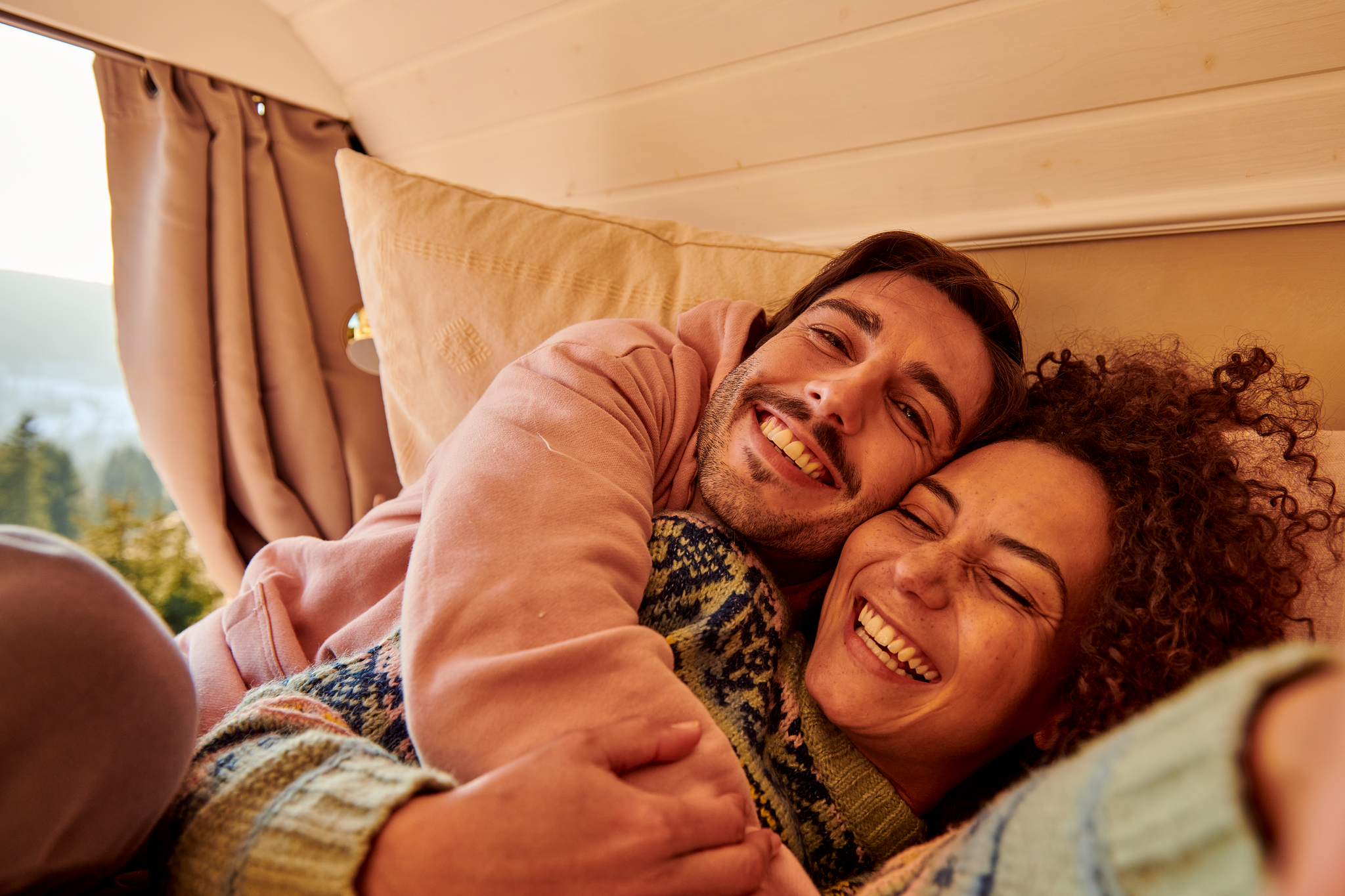 Ein lächelndes Paar kuschelt sich in einem gemütlichen Raum mit holzverkleideten Wänden aneinander, während sanftes Licht durch ein Fenster fällt. 