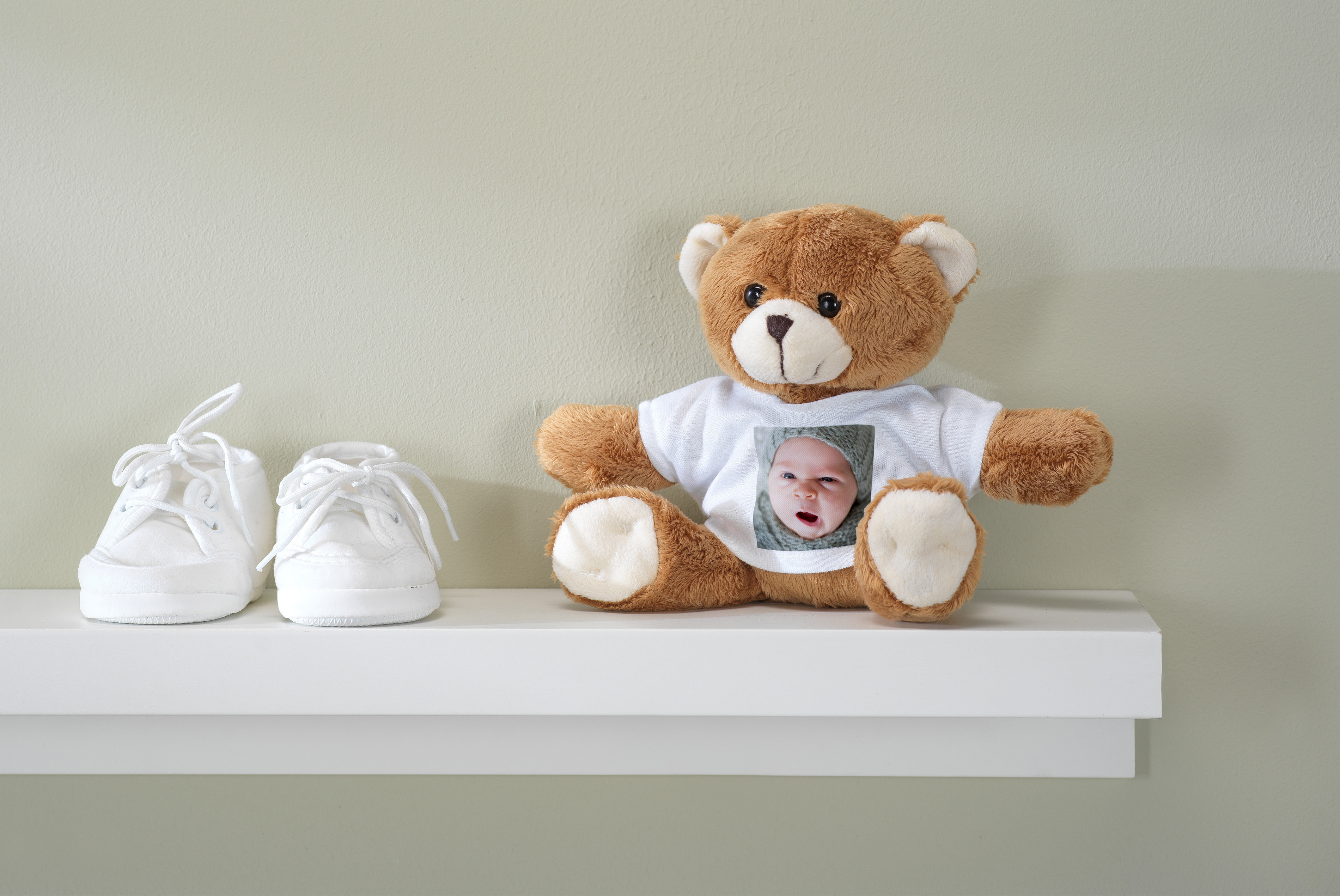 En teddybjørn iført en t-shirt med et billede af en baby sidder på en hvid hylde ved siden af et par små hvide babysko.