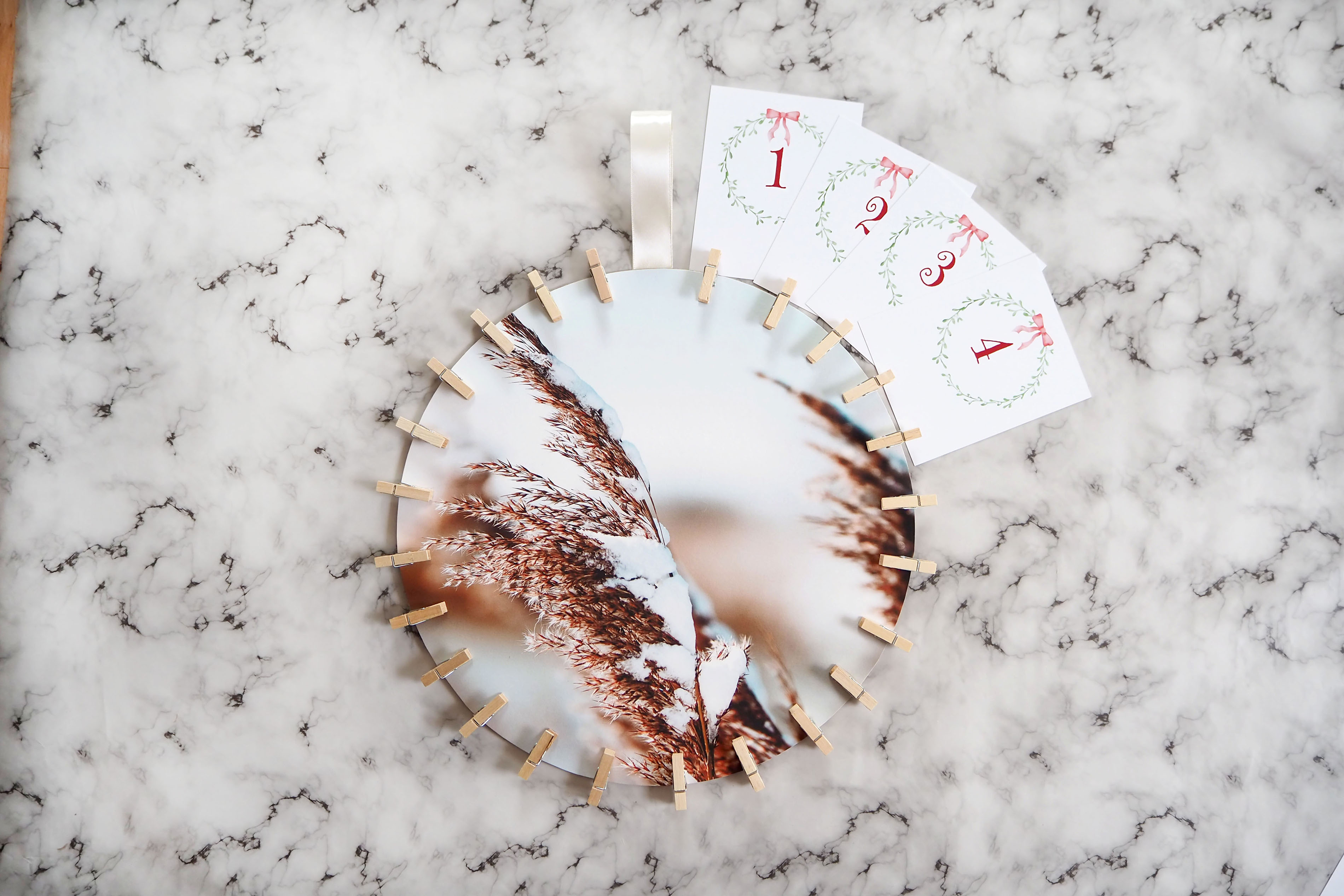 Ein rundes Foto mit schneebedeckten Gräsern liegt auf einem marmorierten Hintergrund. Mit Wäscheklammern sind 4 Karten eines Adventskalenders am Bild befestigt.