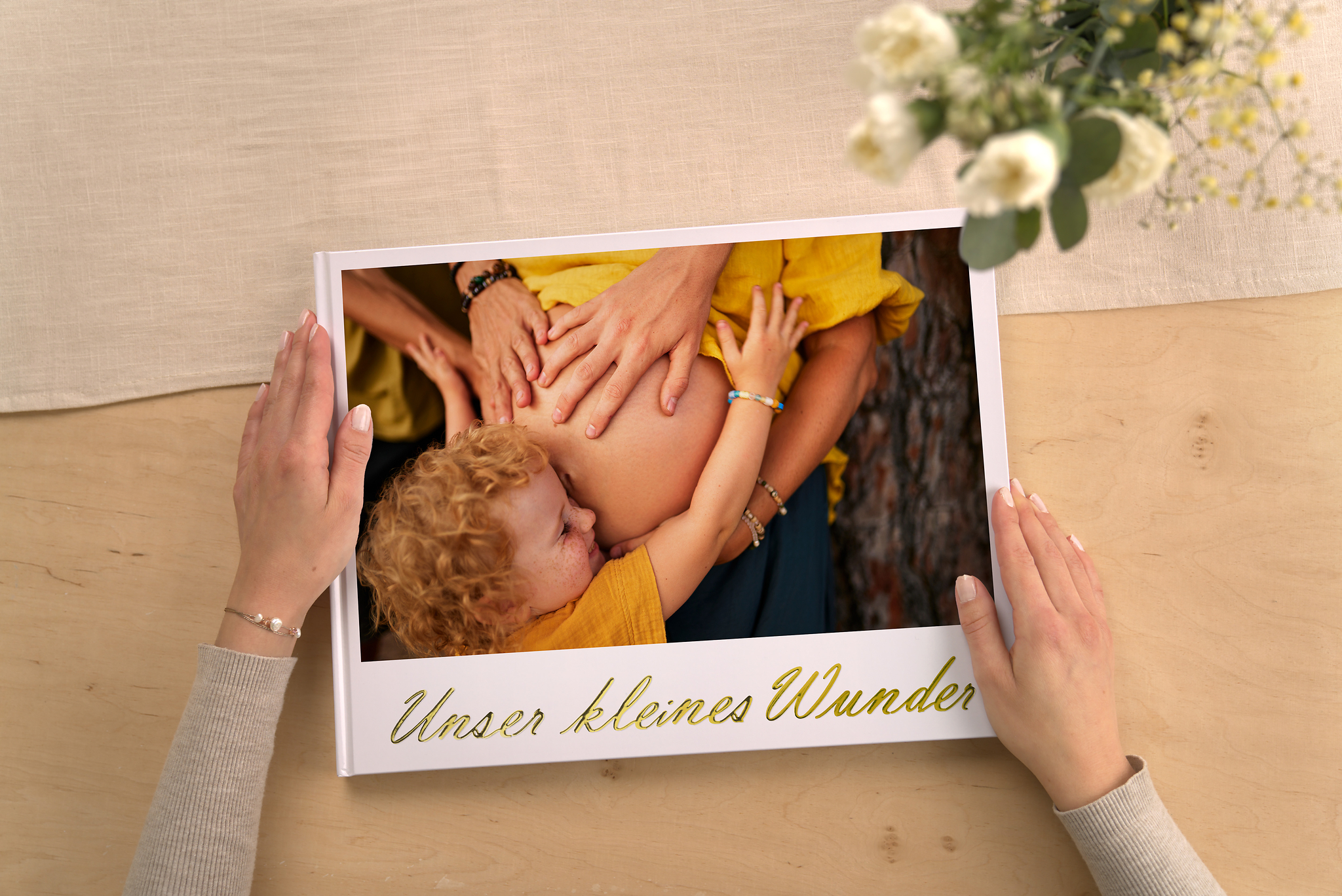 Eine Frau hält ein Fotobuch mit dem Titel "Unser kleines Wunder" in der Hand. Der Titel ist in goldener Schrift veredelt und auf dem Cover ist ein Foto aus der Schwangerschaft zu sehen.