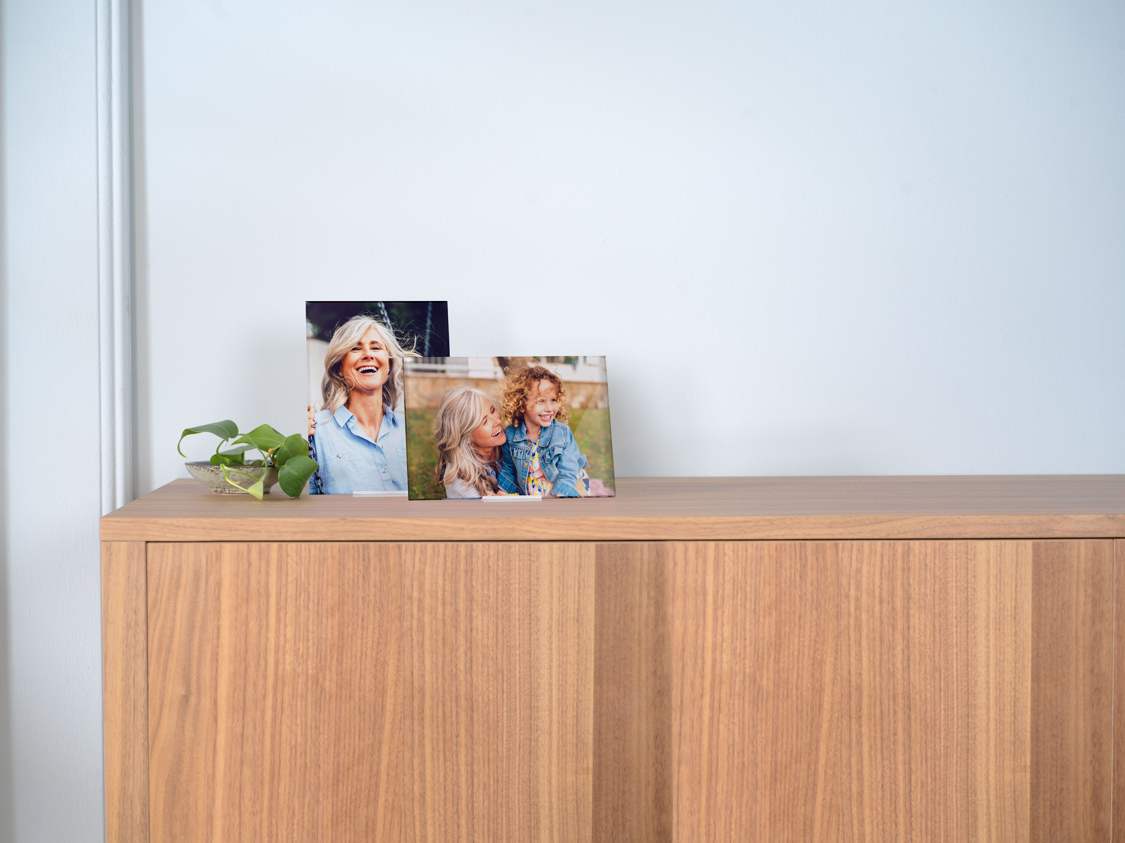 Zwei gerahmte Fotos stehen auf einem hölzernen Sideboard; die Bilder zeigen lächelnde Personen in einer entspannten und fröhlichen Stimmung. 