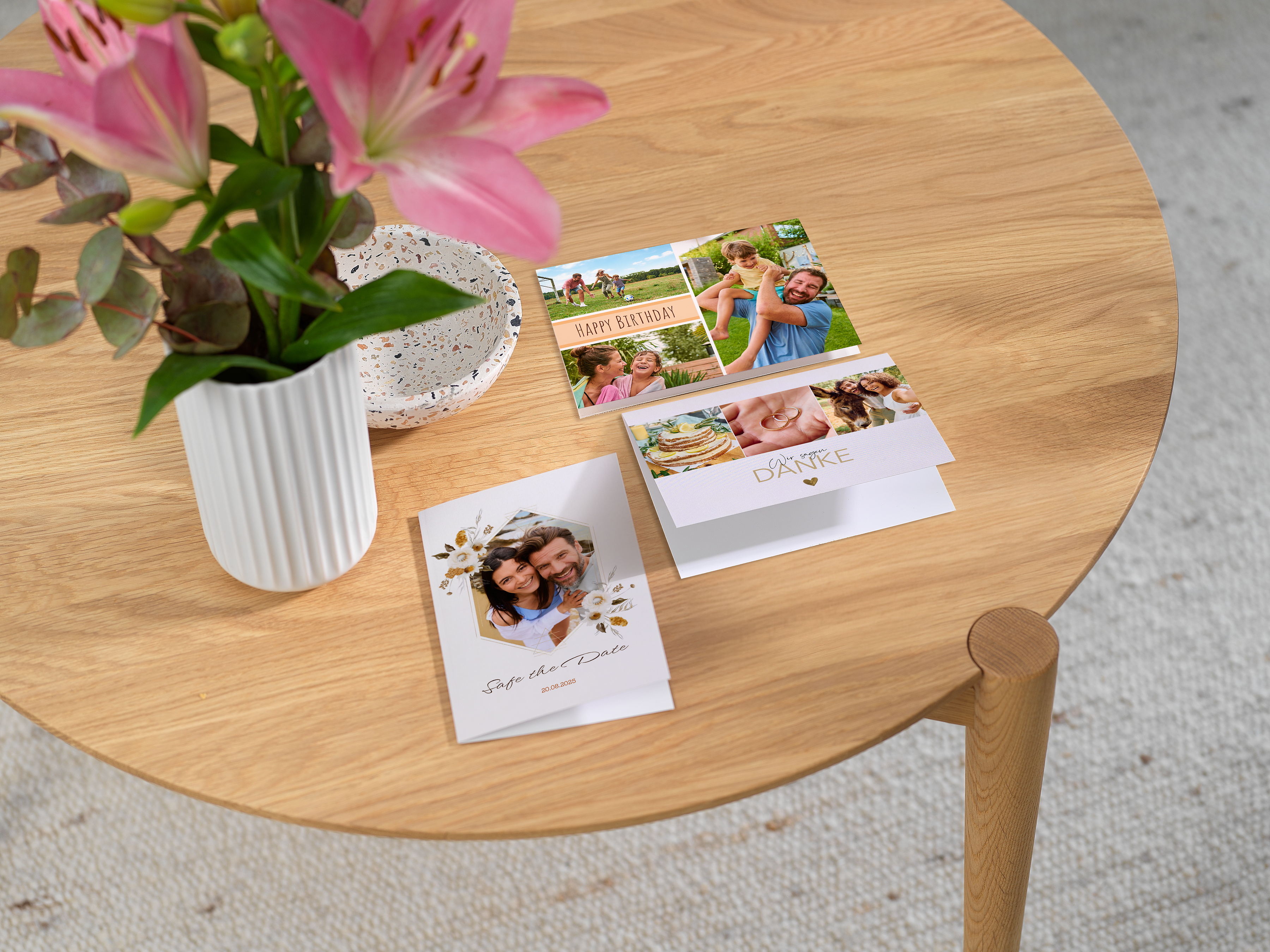 Auf einem runden Holztisch liegen verschiedene individuell gestaltete Fotokarten, darunter eine Geburtstagskarte, eine Dankeskarte und eine Einladung. Neben den Karten steht eine Vase.