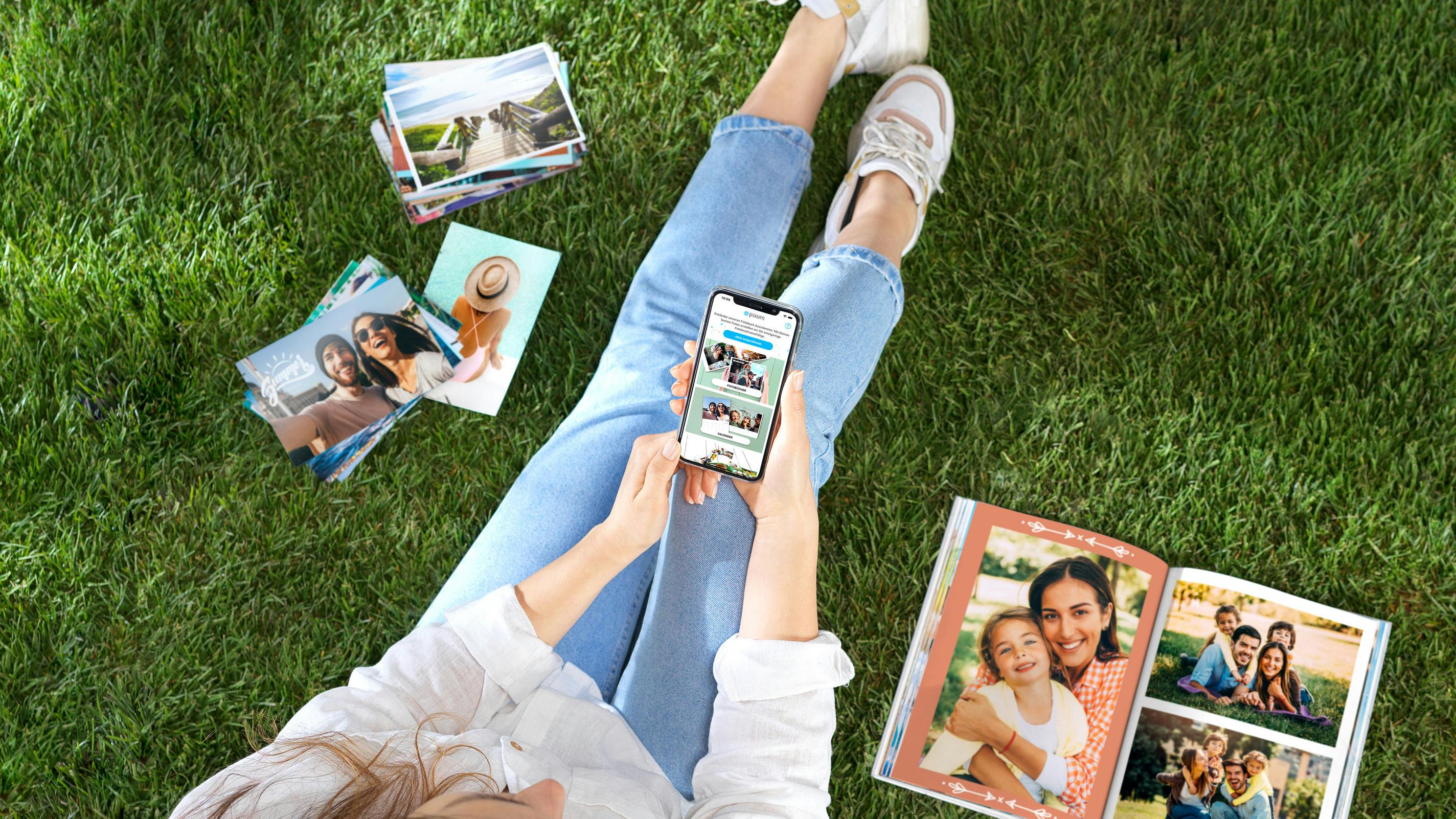Fotoprodukte mit der App bestellen Von oben fotografierte Frau mit einem Handy in der Hand, die auf einer Wiese sitzt und Fotoprodukte gestaltet