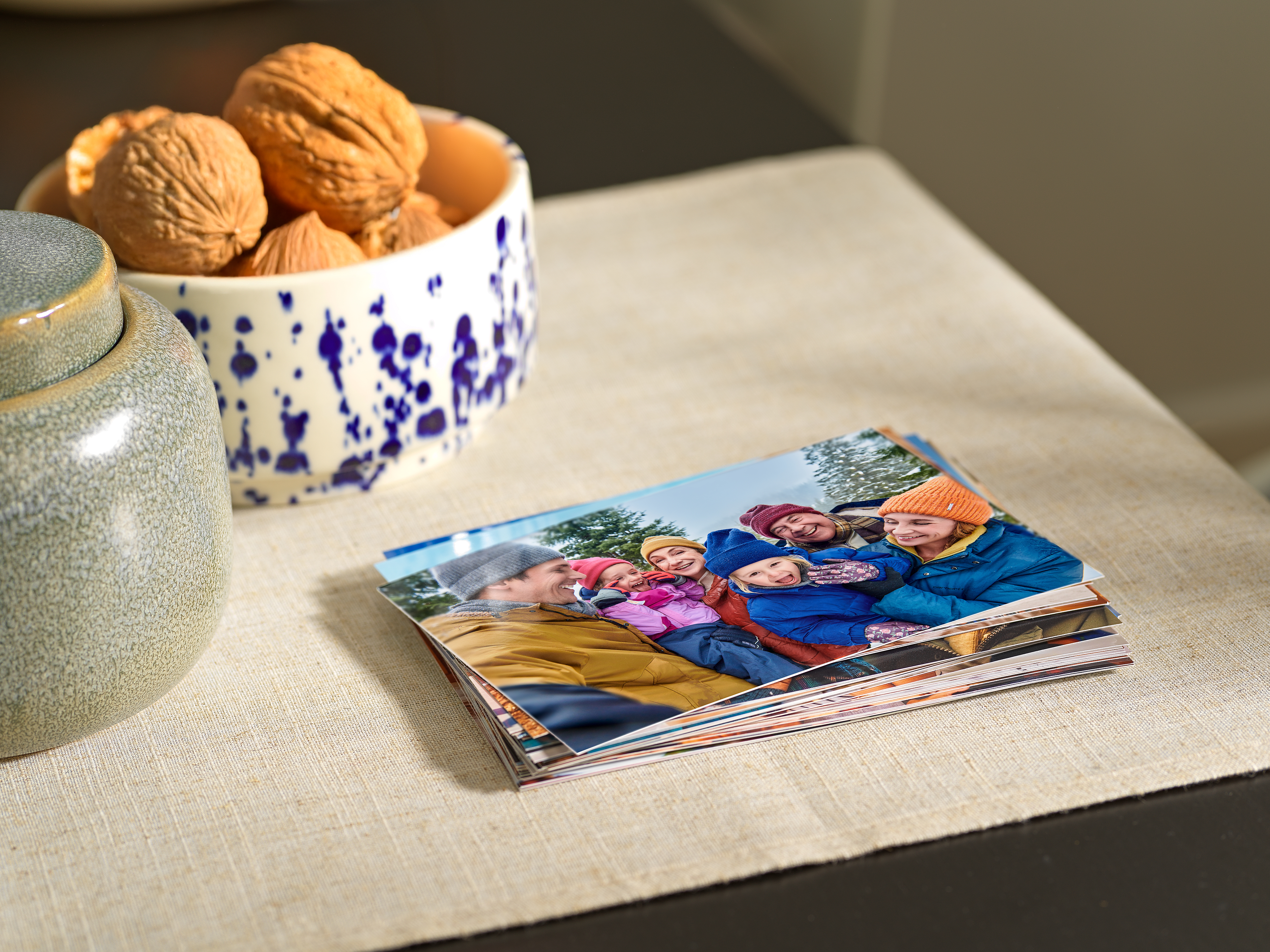 Ein Stapel von Fotoabzügen liegt auf einer beigen Couch. Auf den Abzügen sind Familienfotos abgebildet.