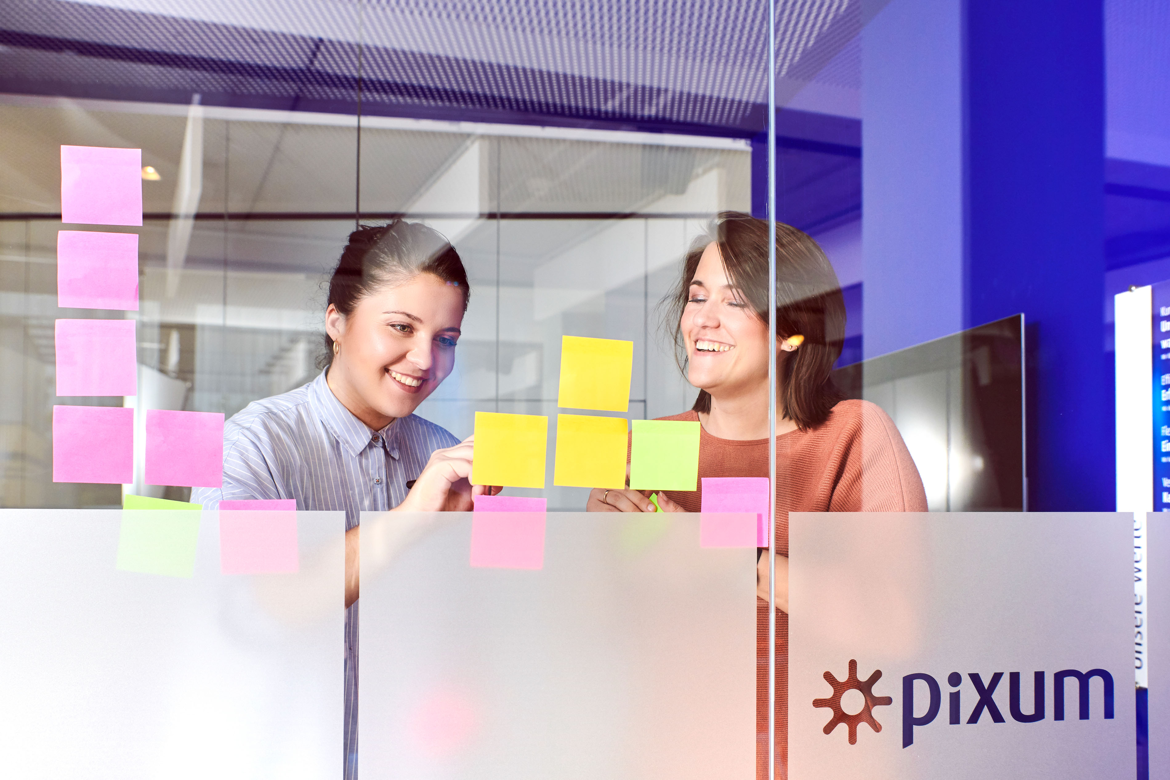Pixum Mitarbeitende in einem Meetingraum, die Post-its an einer Glaswand beschriften